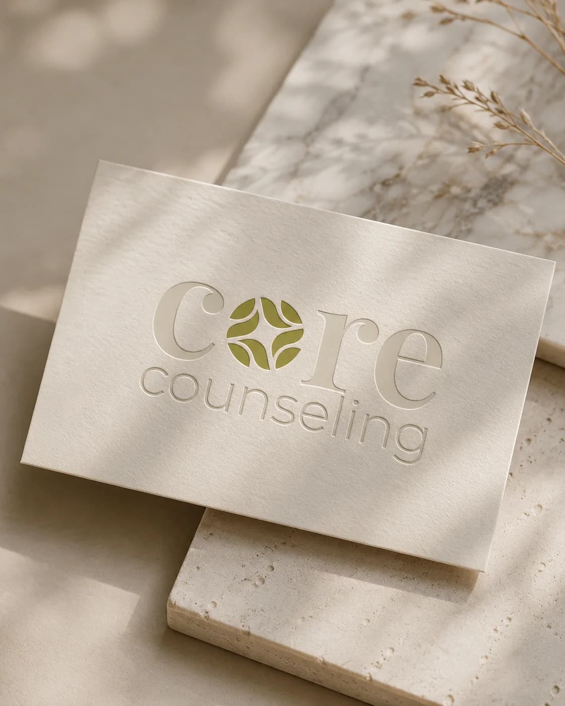 Letterpressed business card with the Core Counseling logo debossed in cream and olive, resting on travertine in dappled light